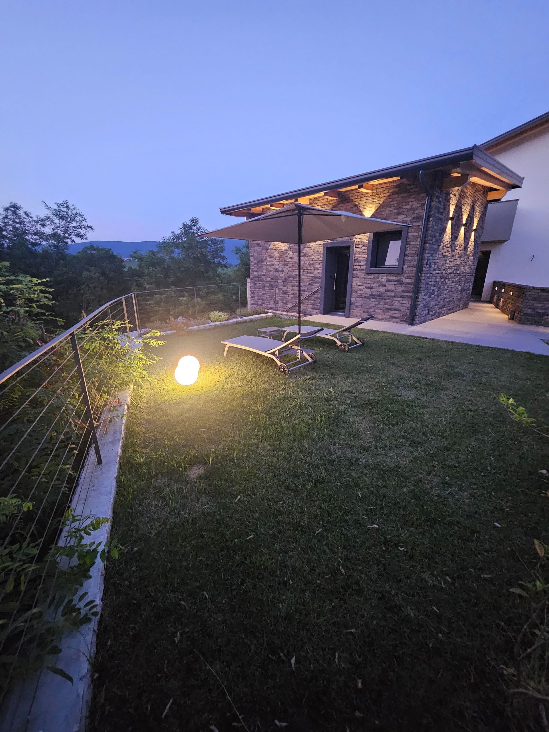 Il patio di una casa in pietra al crepuscolo, con il prato illuminato, le sedie a sdraio e la vista sulla collina sotto un cielo azzurro.