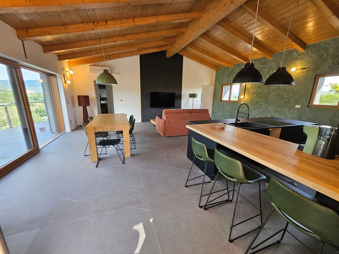 Zona pranzo in stile loft moderno con travi in ​​legno a vista, lungo tavolo, sgabelli da bar verdi, camino e ampie finestre.