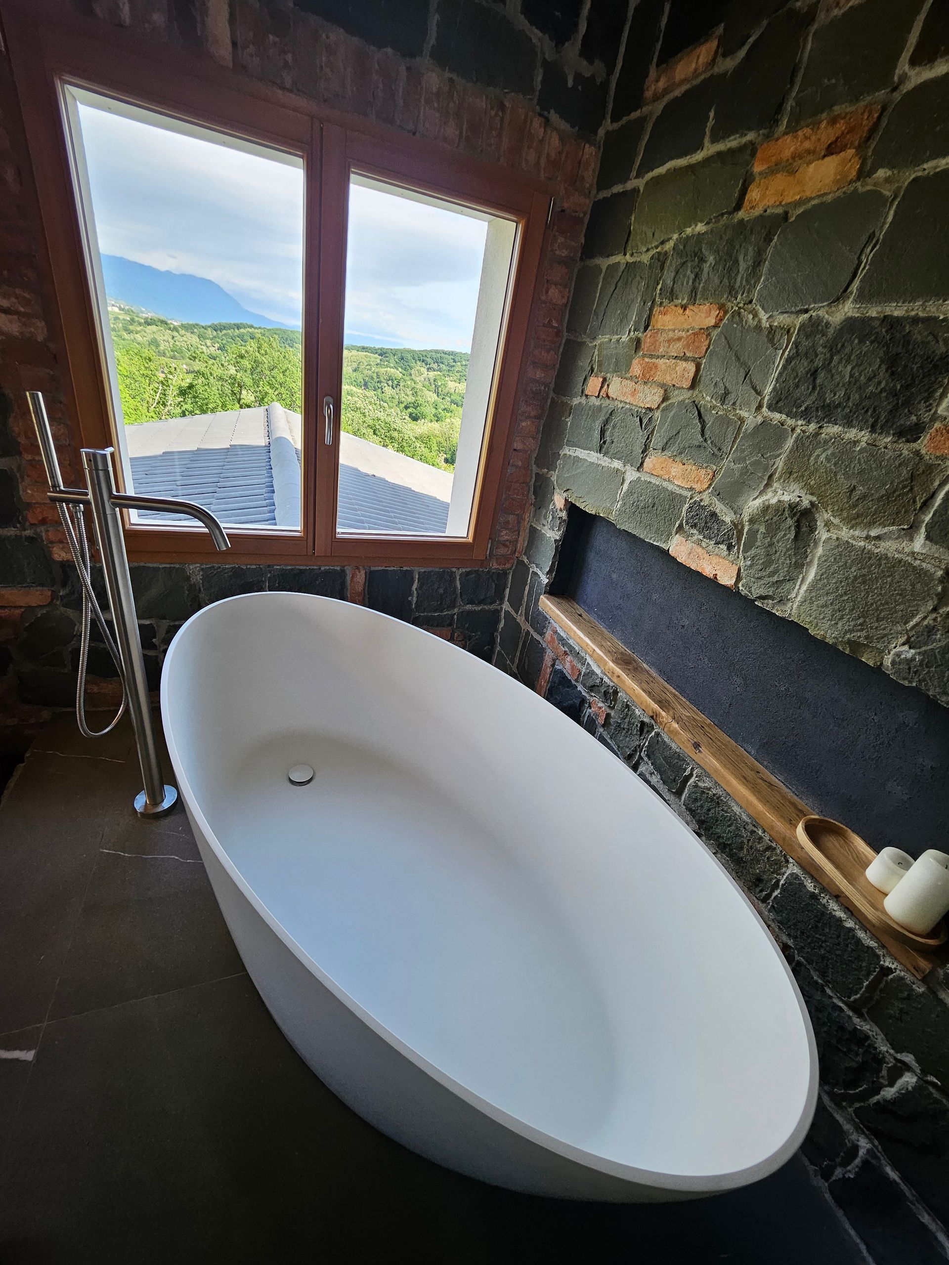 Vasca da bagno bianca freestanding accanto a una finestra con vista sulle montagne in un bagno con pareti in pietra.
