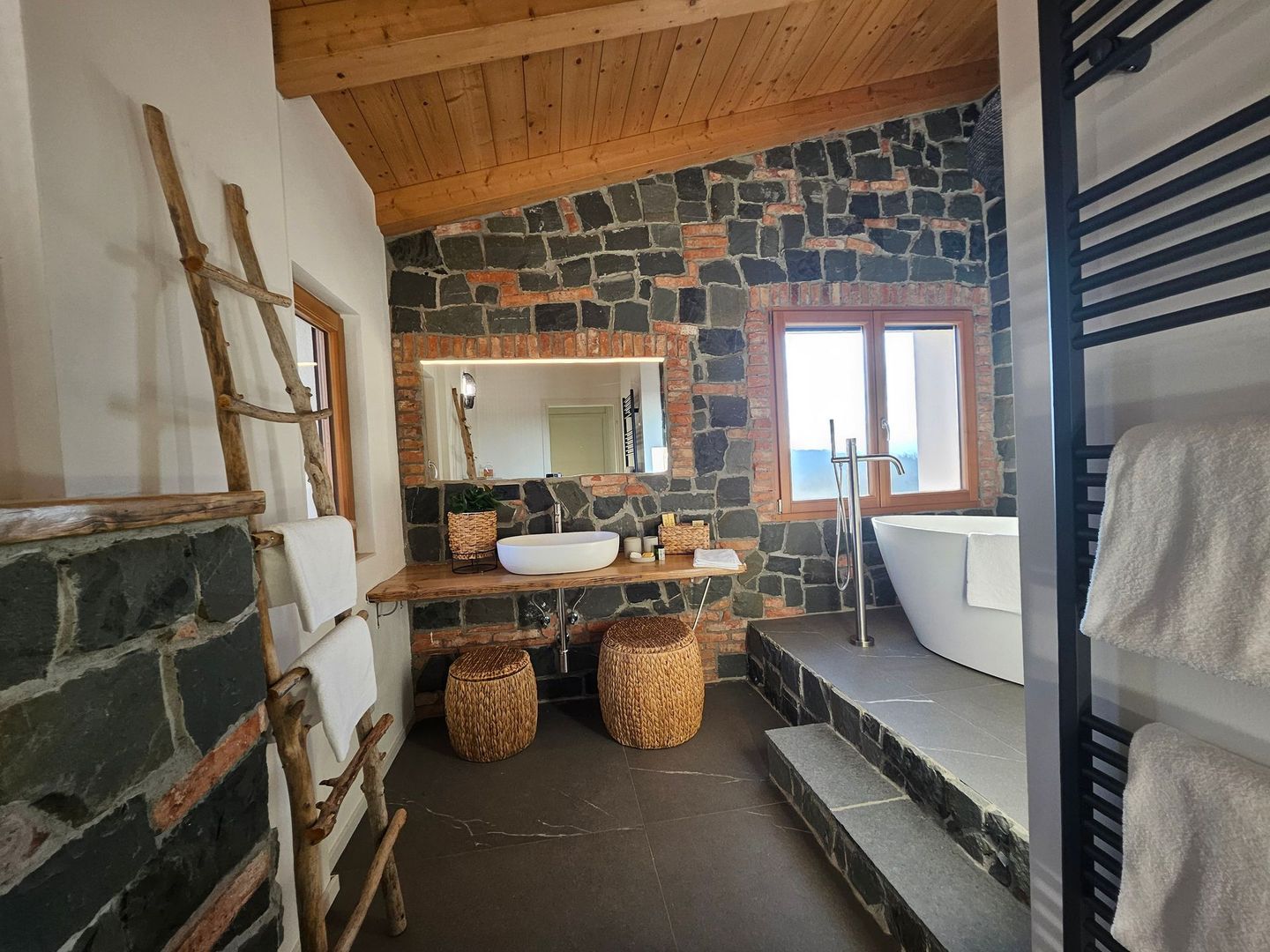 Bagno rustico con pareti in pietra, soffitto in legno, sgabelli rotondi in vimini, doppio lavabo, vasca e mensole a scala.