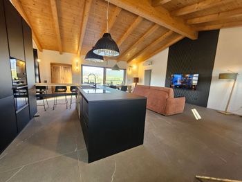 Soggiorno moderno a pianta aperta con soffitto in legno, isola cucina nera e divano rosa accanto a grandi finestre.