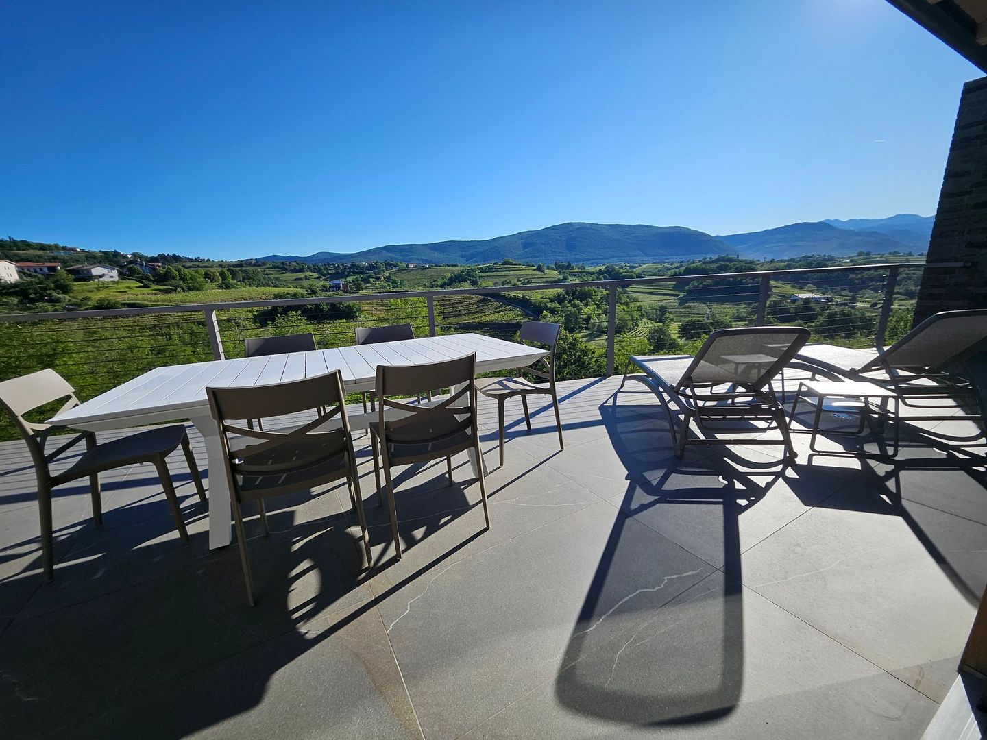 Patio esterno con tavoli e sedie che si affaccia su una soleggiata vallata verde e sulle colline in lontananza.