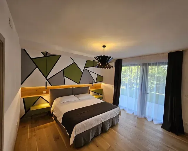 Camera da letto moderna con decorazioni murali geometriche, letto king size, illuminazione calda e ampia finestra con tende.