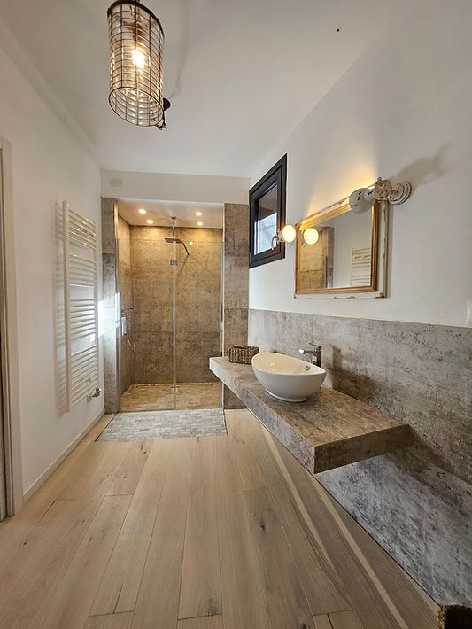 Luminoso bagno moderno con cabina doccia, lavabo sospeso, specchio e finiture in legno chiaro e pietra.