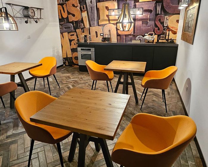 Interno di un piccolo caffè con tavoli in legno e sedie arancioni, addossato a una parete decorata con graffiti.