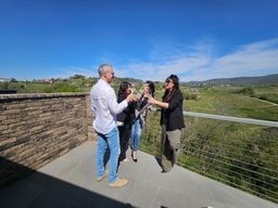 Tre persone si salutano su una terrazza in pietra con vista sulle montagne, sotto un cielo azzurro.