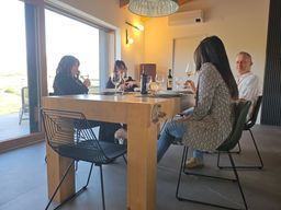 Persone sedute attorno a un tavolo da pranzo in legno in una stanza moderna e luminosa, con una porta aperta verso l'esterno.