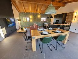 Moderna zona pranzo e cucina a pianta aperta con soffitto in legno, sedie verdi e divano scuro vicino al televisore.