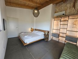 Camera da letto moderna con soffitto in legno, letto bianco, pavimento in piastrelle scure e mensole a vista sulla destra.