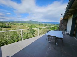 Patio con tavolo e sedie affacciato su una rigogliosa valle verde e sulle montagne in lontananza, sotto un cielo azzurro.