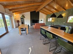 Soggiorno moderno con soffitto a volta in legno, moquette grigia, tavolo da pranzo, divano e isola cucina con sgabelli da bar.