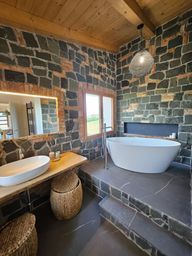 Bagno rustico con pareti in pietra, soffitto in legno, vasca freestanding, lavabo a ciotola e cesti intrecciati.