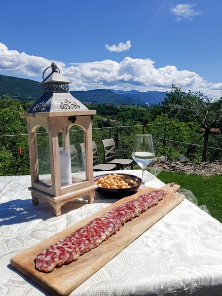 Tavolo all'aperto con carne alla griglia, calice di vino, lanterna e vista sulle montagne sotto un cielo azzurro.