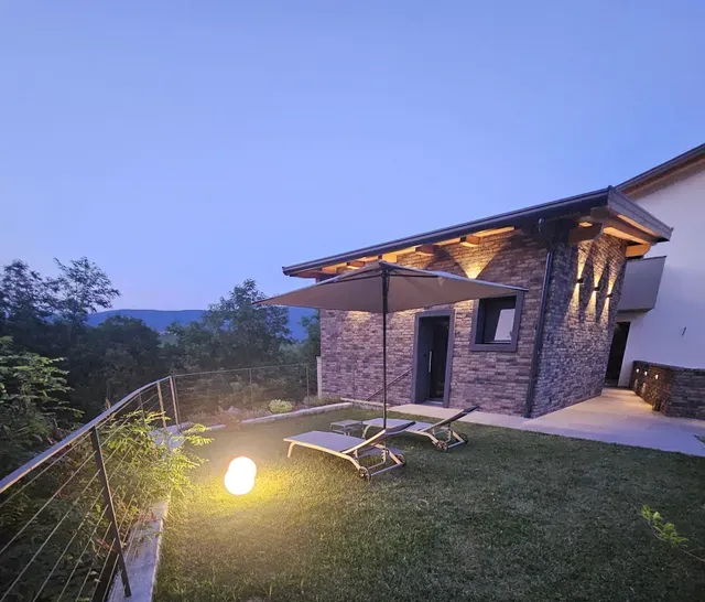 Moderna baita in pietra con patio illuminato e sedie a sdraio al crepuscolo, affacciata su una collina erbosa.