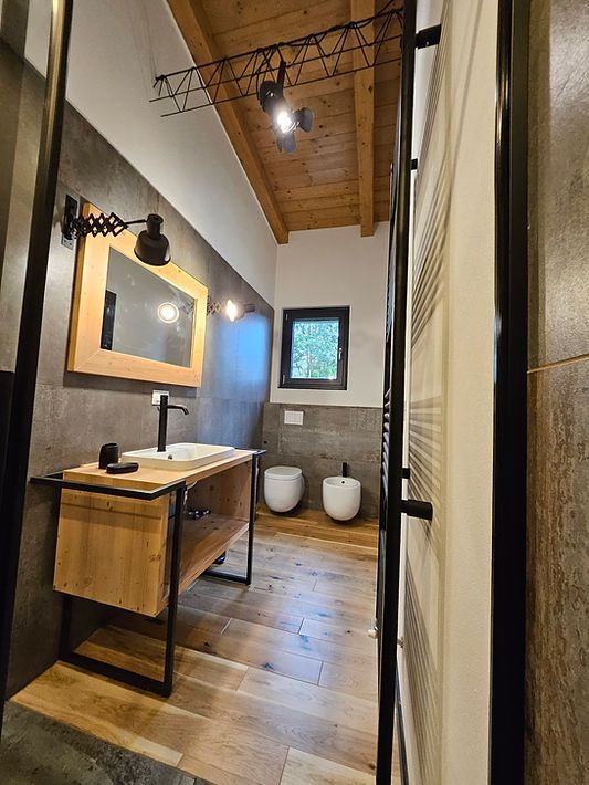 Bagno moderno con mobile lavabo in legno, due WC bianchi e pareti piastrellate sotto un soffitto in legno.