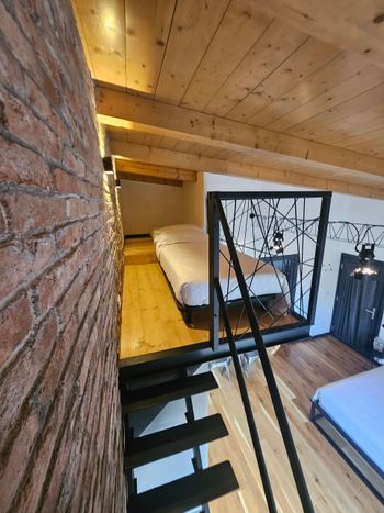 Camera da letto soppalcata con due letti singoli, ringhiera in metallo nero, soffitto in legno e parete in mattoni.