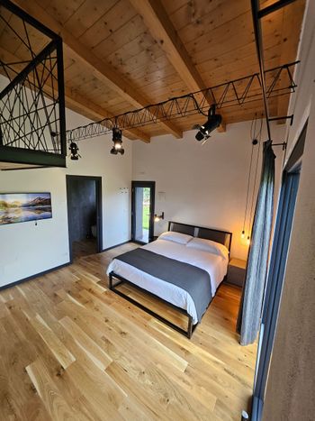 Camera da letto soppalcata con letto, pavimento in legno, soffitto con travi a vista e dettagli in metallo nero.