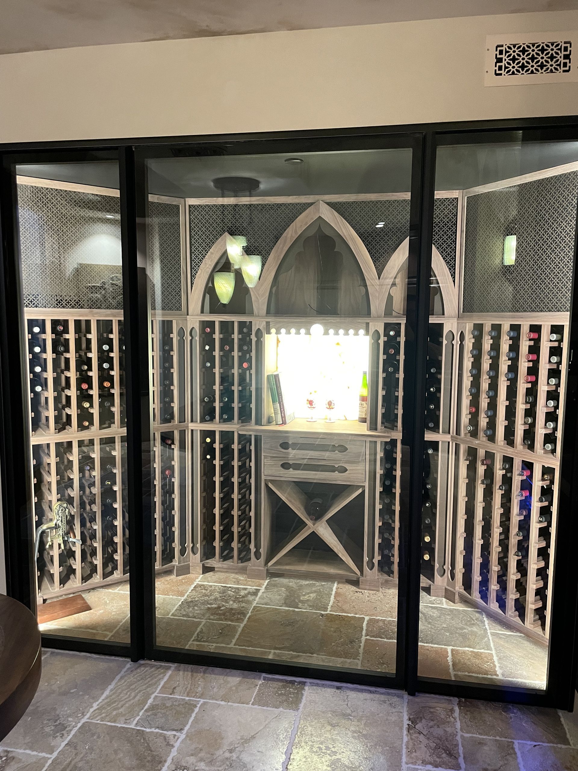 Glass wine cellar with black metal framing and arched doorway, lit shelves and stone tile floor