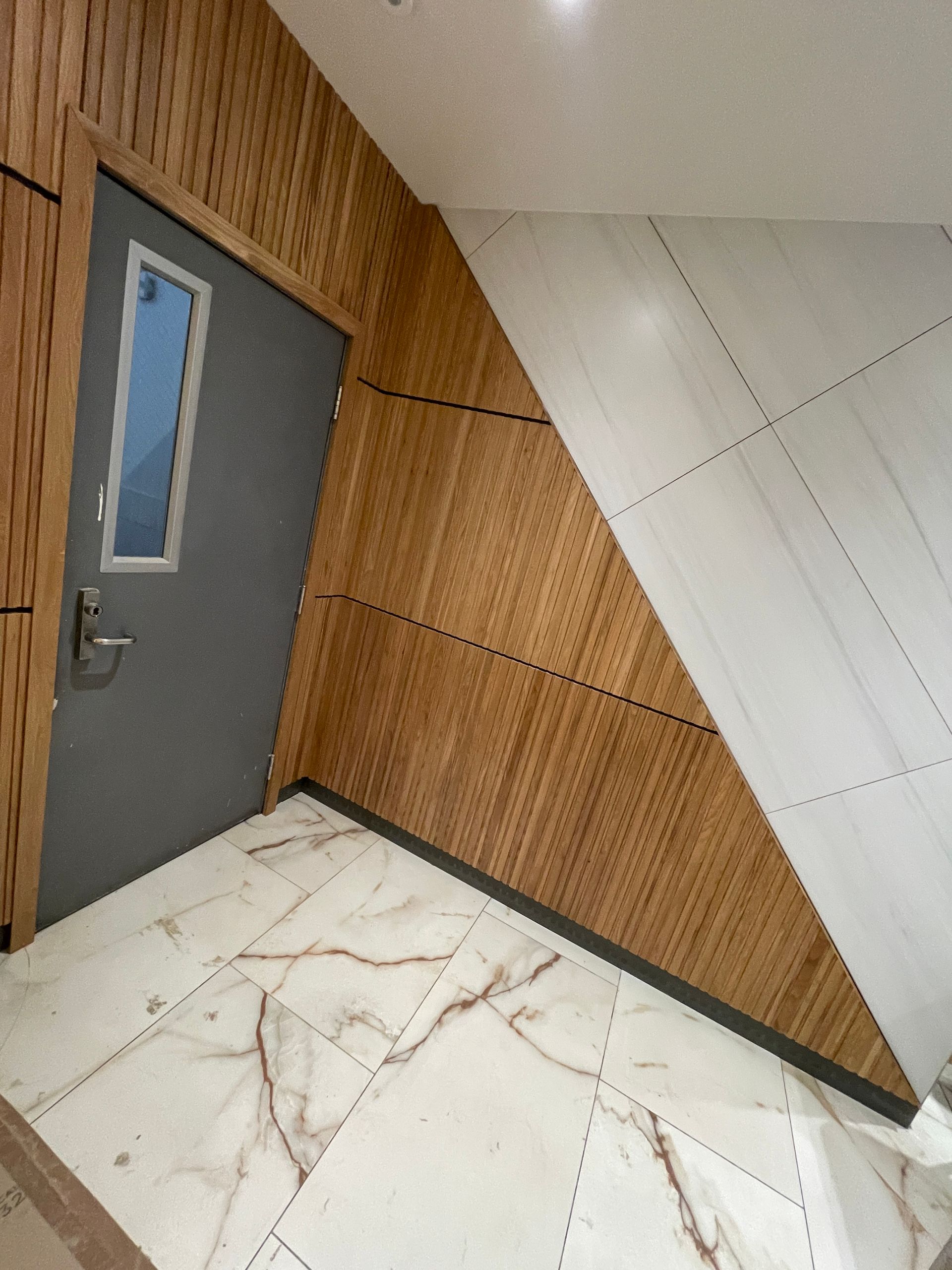 Modern lobby corner with a gray door, wood-paneled walls, and white marble floor.
