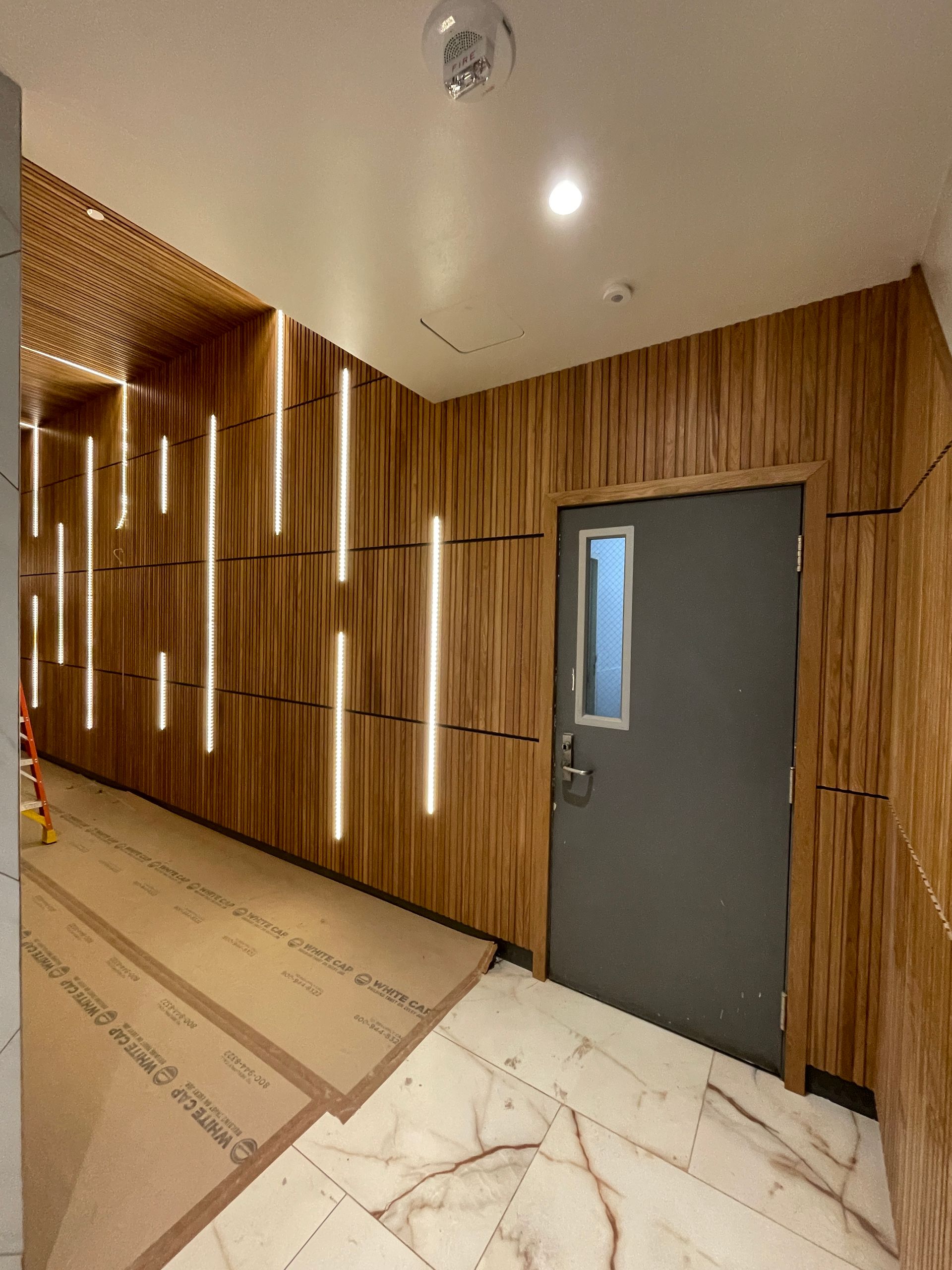 Wood-paneled hallway with a gray door, white marble floor tiles, and recessed ceiling lights.