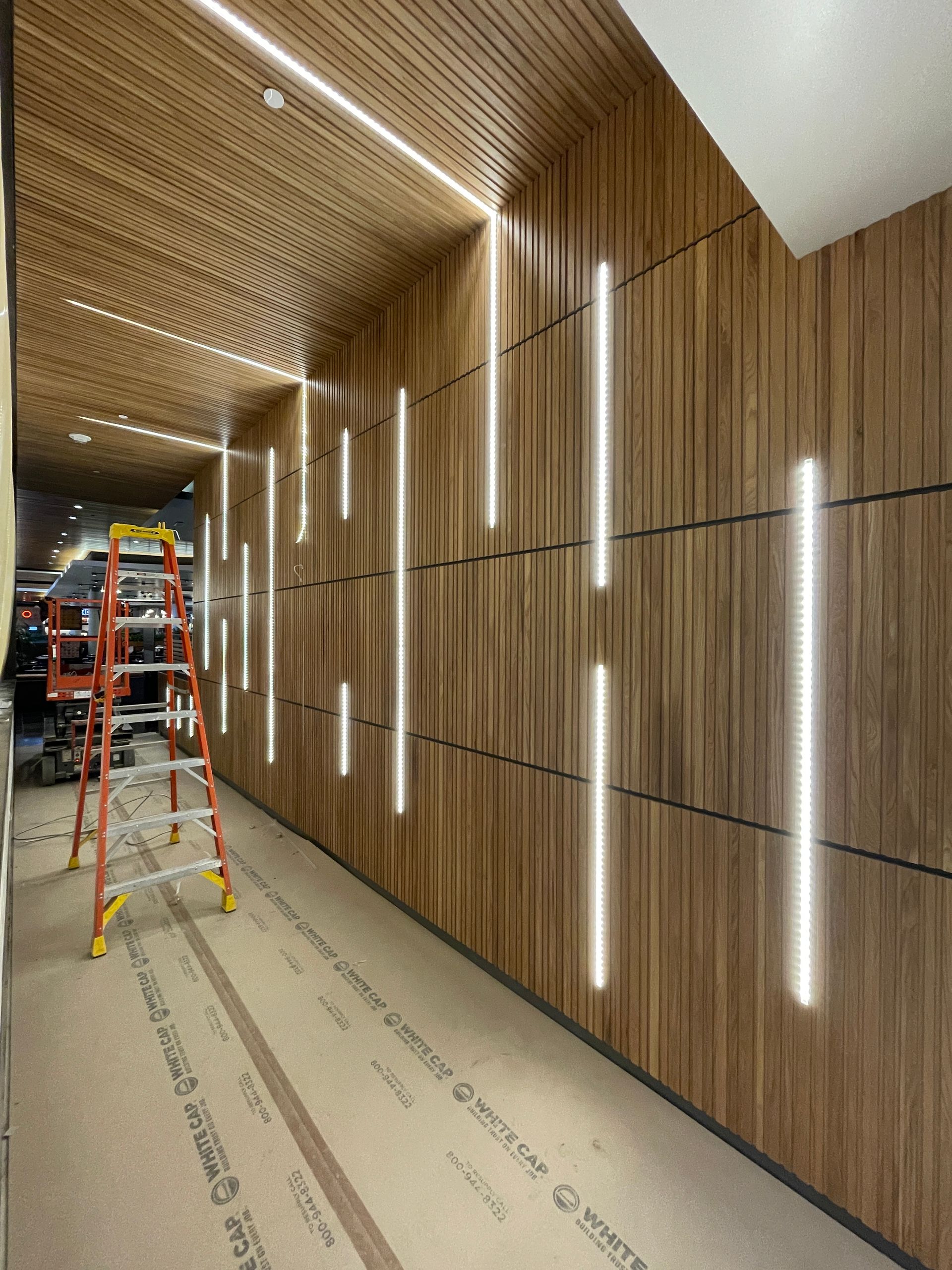 Long hallway under construction with wood-paneled walls, vertical lights, and ladders on a covered floor