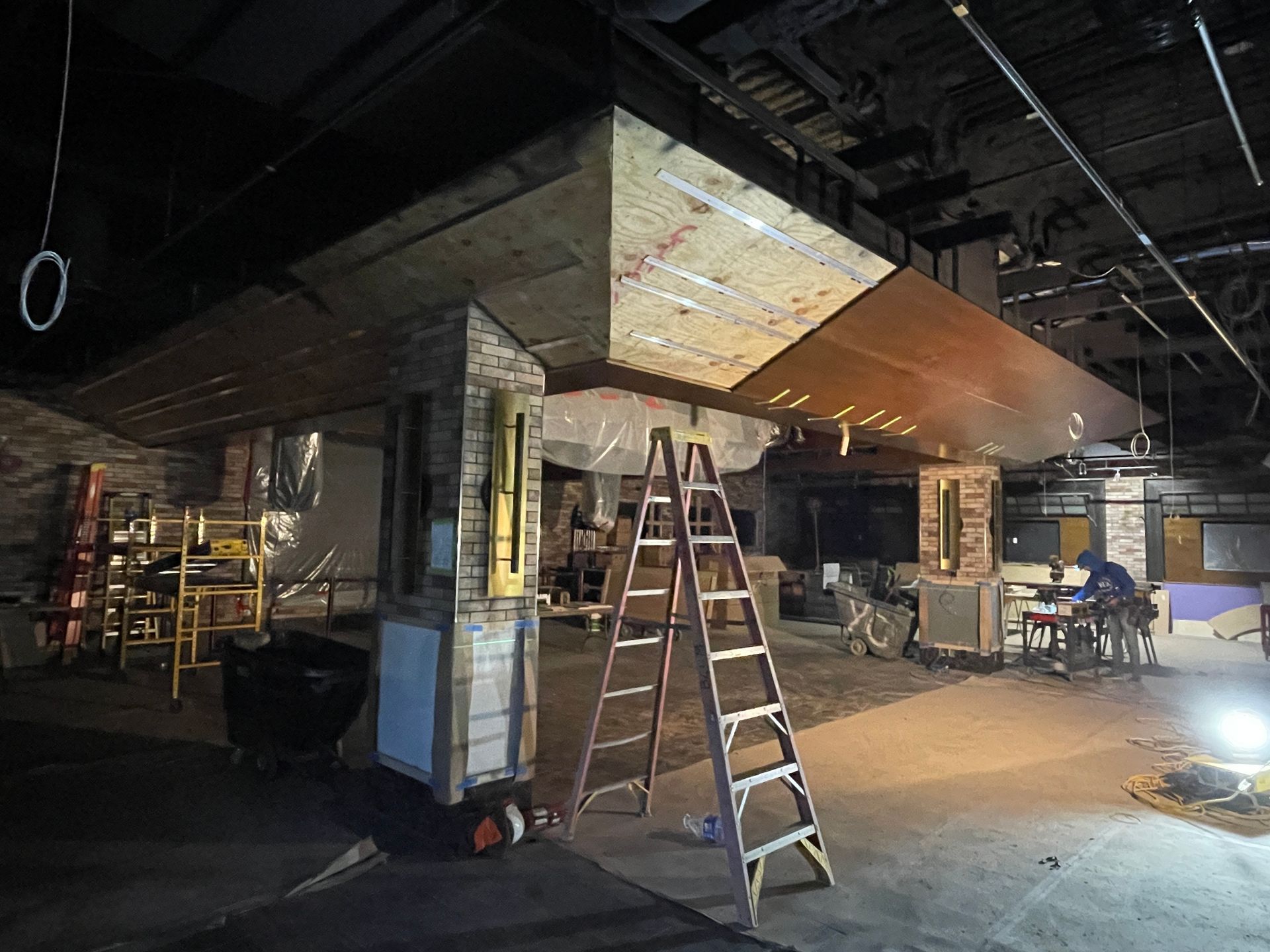 Dim, unfinished room with ladders, construction materials, and overhead ductwork under fluorescent lights