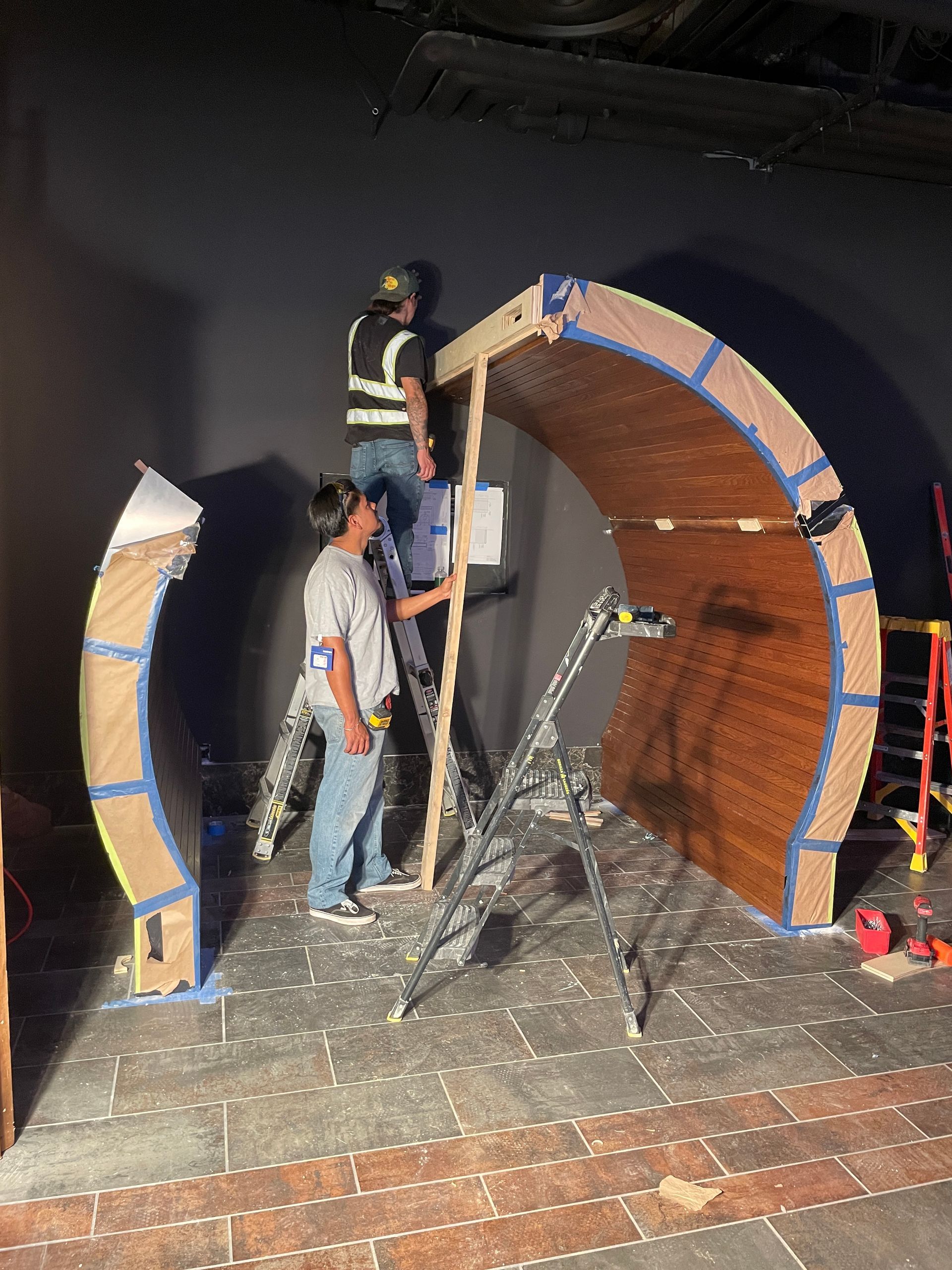 Two people assembling a large curved wooden structure in a dim indoor space