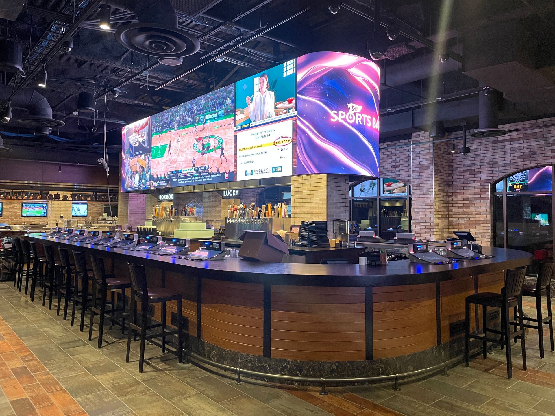 Empty sports bar with a long counter, stools, and large glowing screens on the wall.