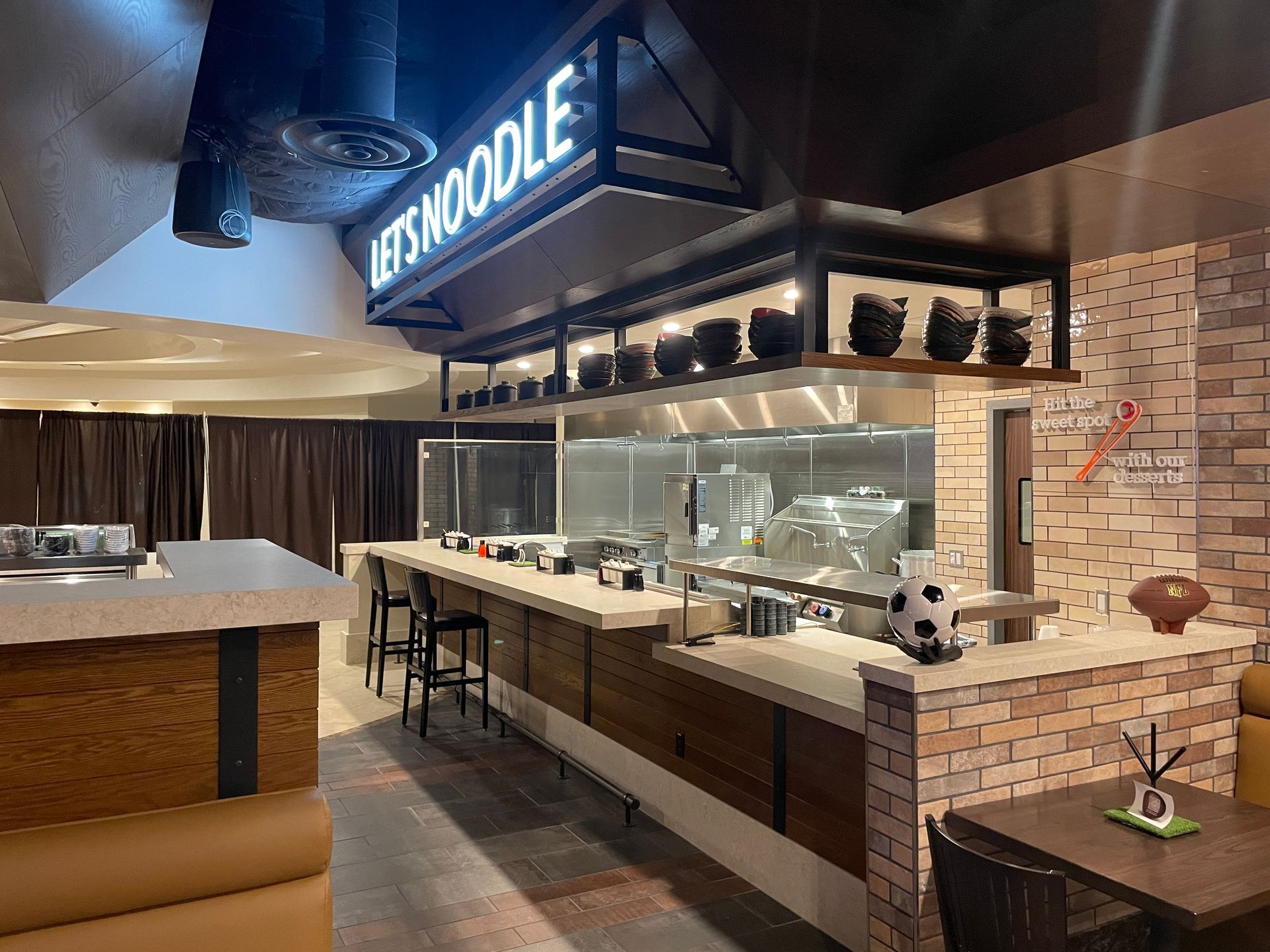 Sushi restaurant counter with glass display, black stools, and a lit “Noodles” sign above.
