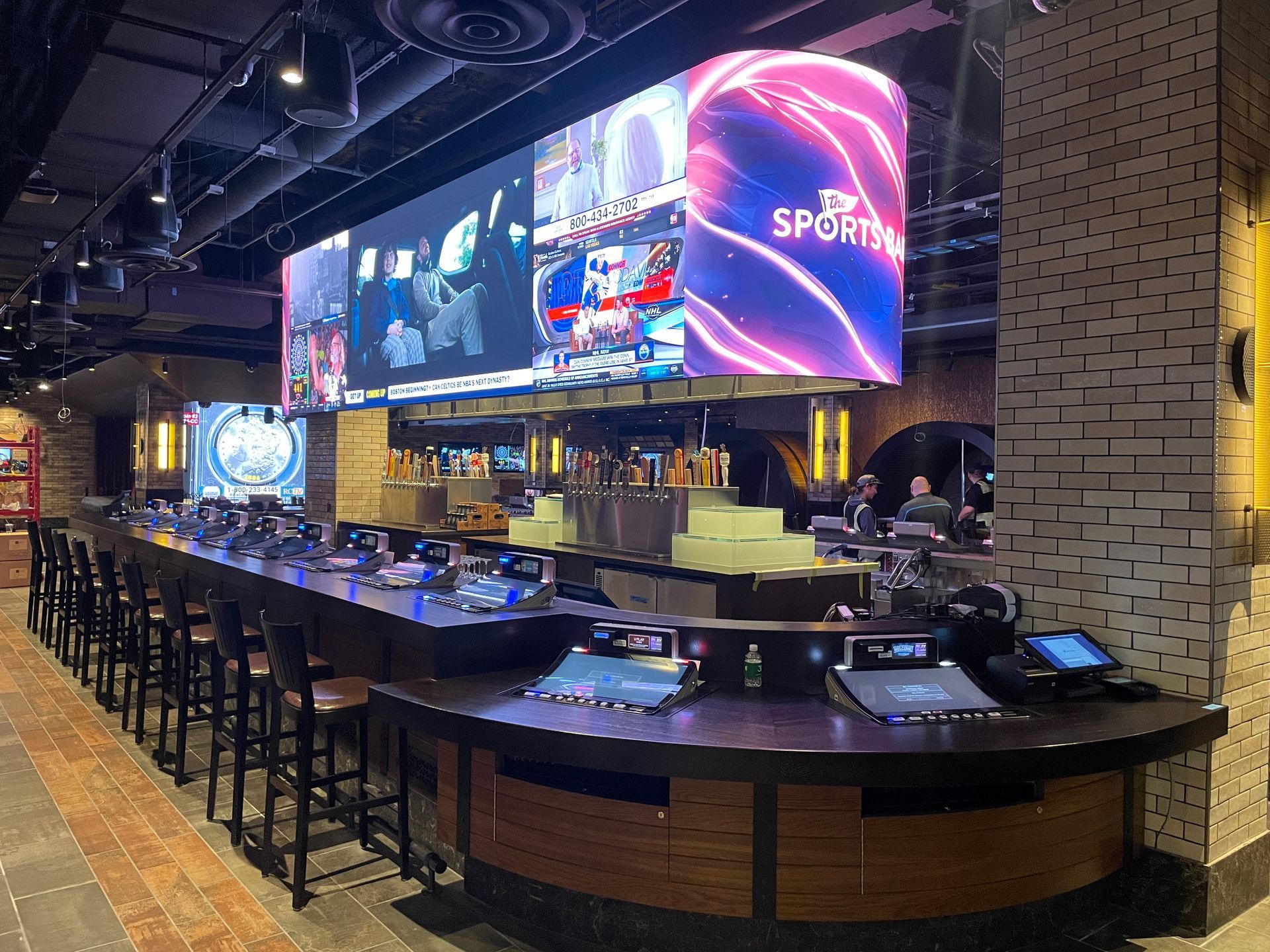 Sports bar interior with a long curved counter, stools, and large glowing TV screens above the bar