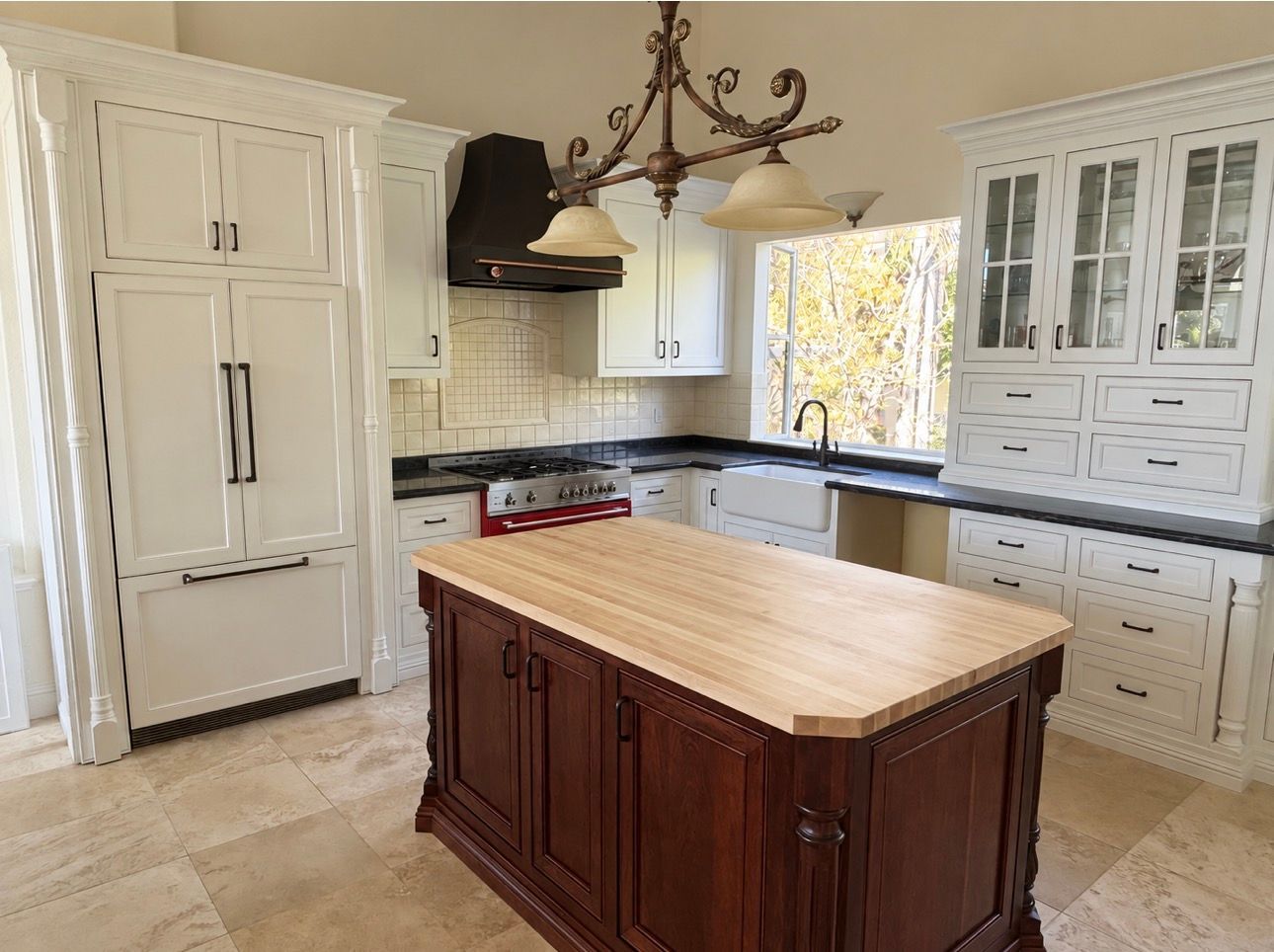 Bright white kitchen with a dark wood island, tiled floor, and large cabinets under a chandelier.