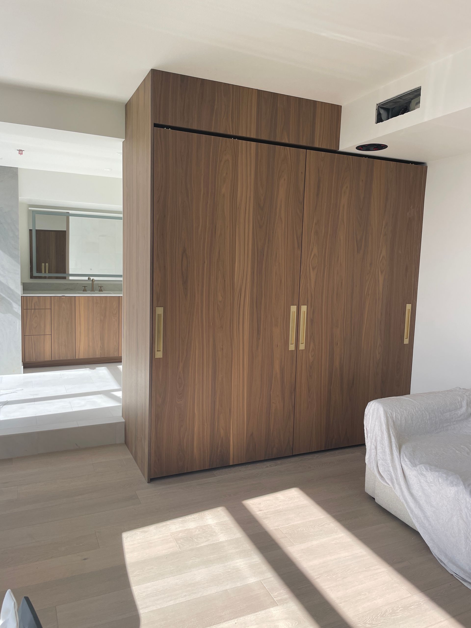 Small bedroom with a large wooden wardrobe, bed edge, and sunlight on the floor near a doorway.
