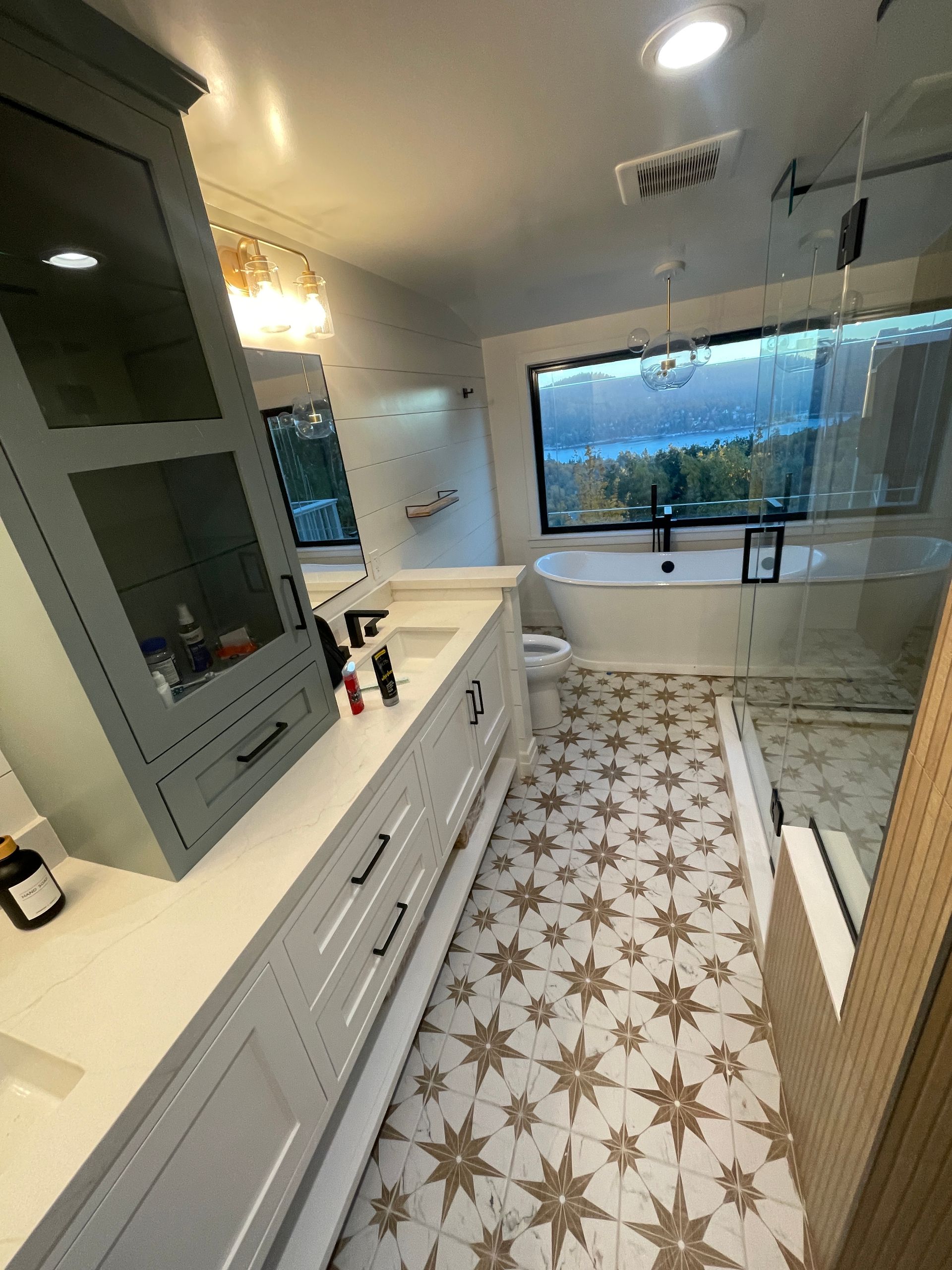 Bright modern bathroom with double vanity, walk-in shower, patterned tile floor, and large window overlooking greenery