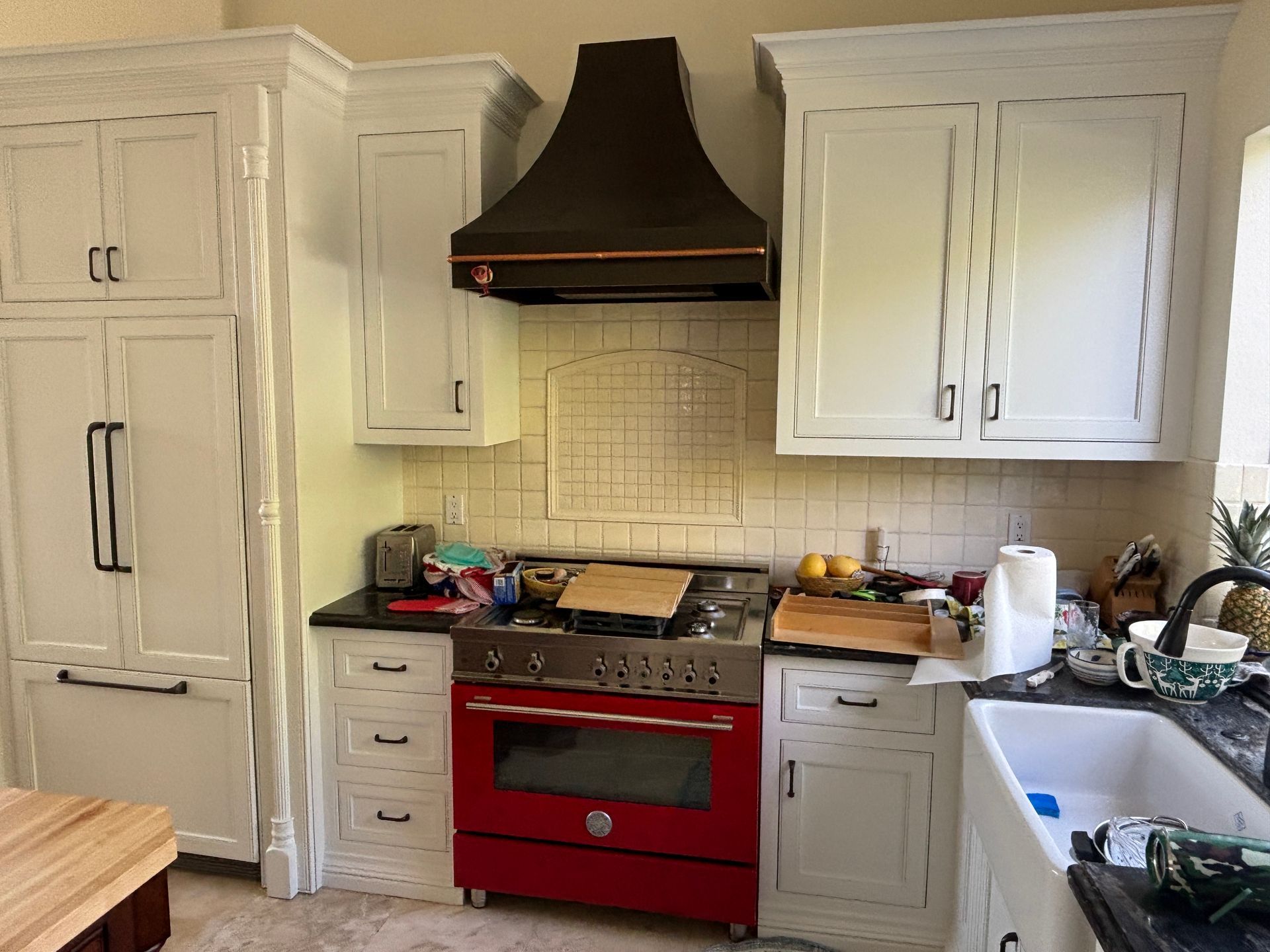 Kitchen with red stove, dark range hood, cream cabinets, and a cluttered countertop by a white sink.