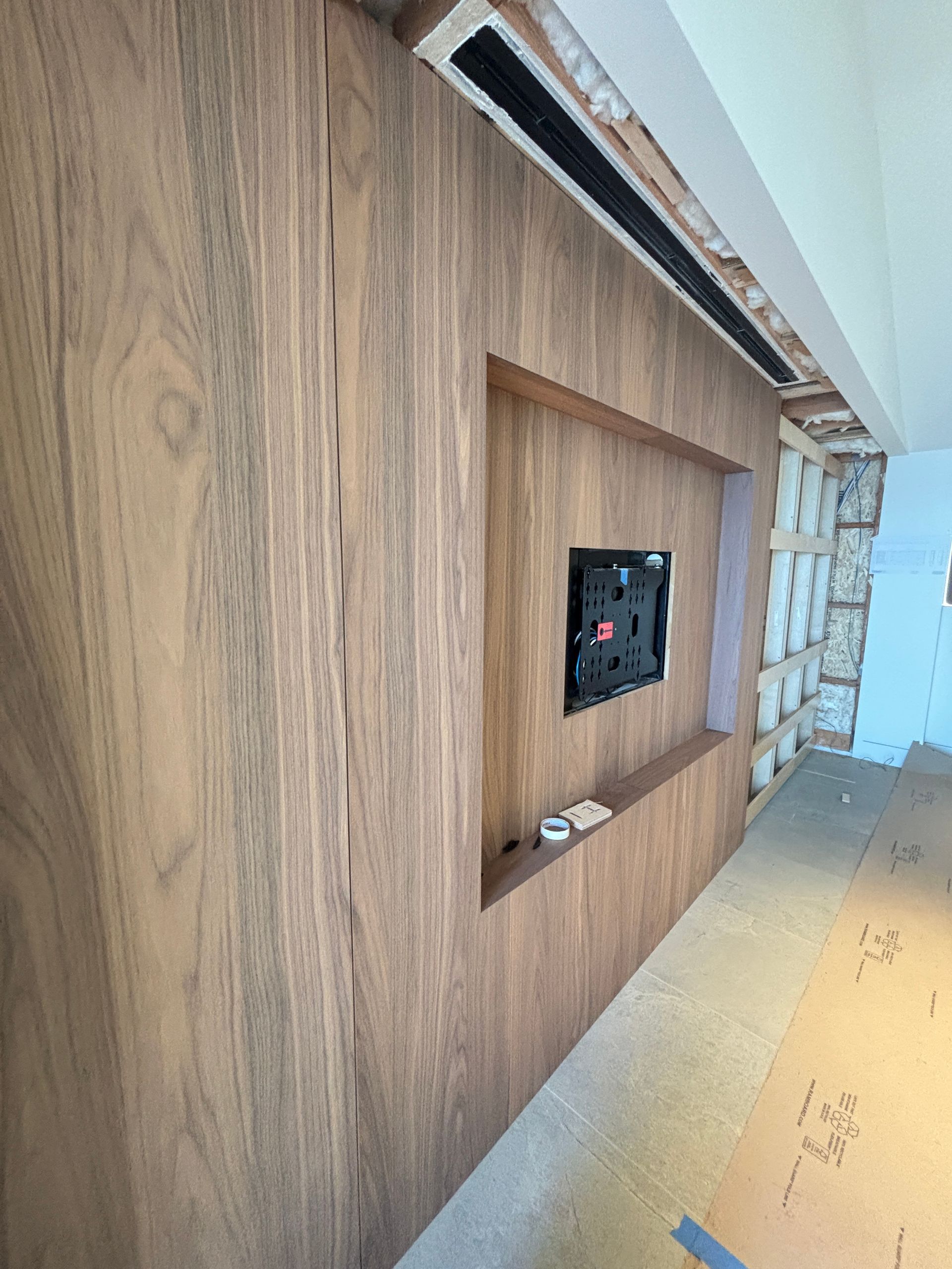 Wood-paneled built-in wall with a recessed black TV mount and exposed ceiling near a hallway under construction