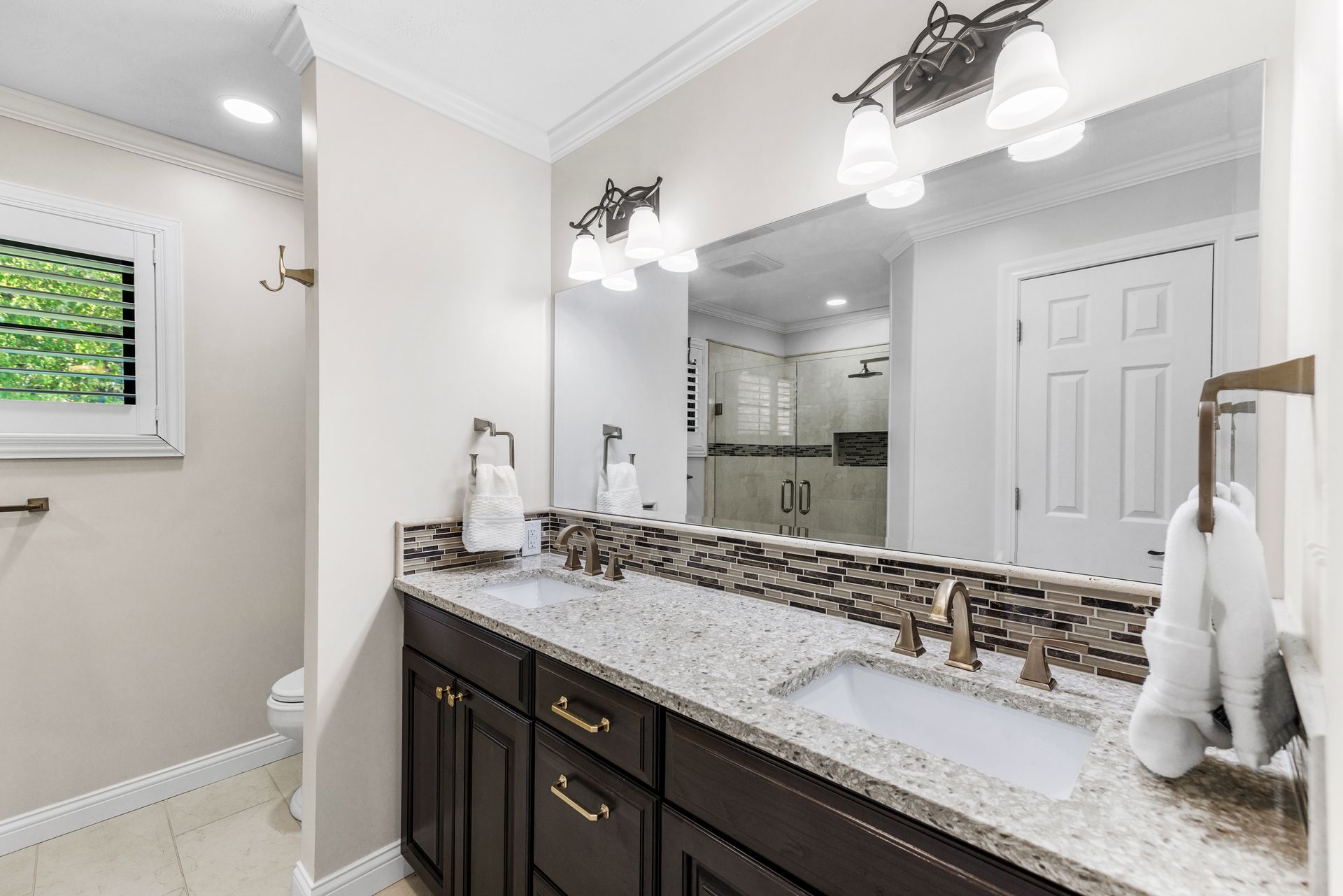 Bathroom with dual sinks, dark brown cabinets, granite countertop, large mirror, and decorative tile backsplash.