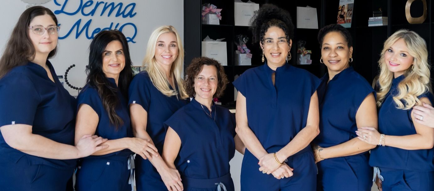 Eight women in blue scrubs pose in a medical office, some with arms crossed. 
