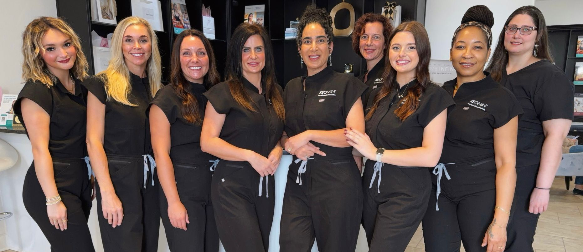 Eight women in blue scrubs pose in a medical office, some with arms crossed.