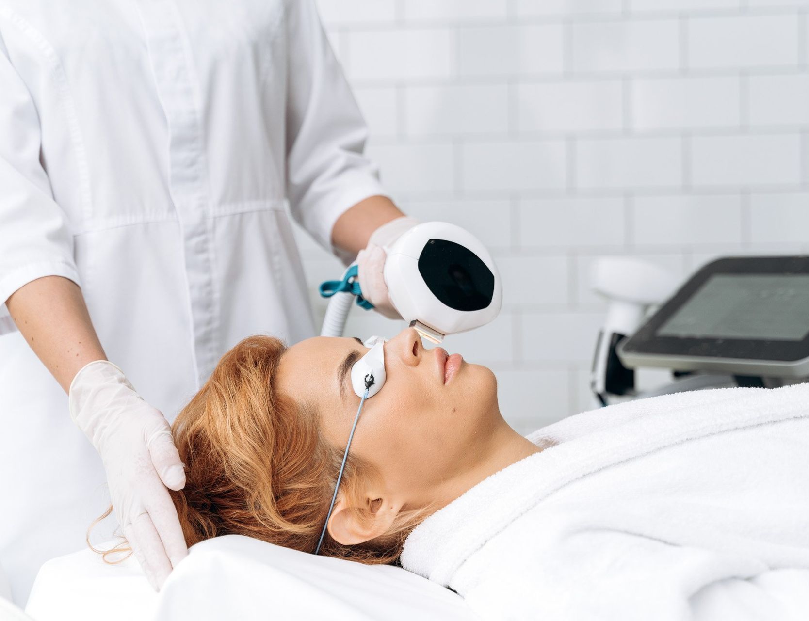Woman receiving facial laser treatment at a spa. She wears protective eye gear while a technician holds the laser device nearby.