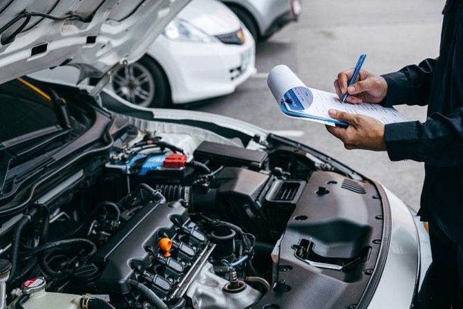 Mechanic inspecting a car engine while writing on a clipboard in a garage