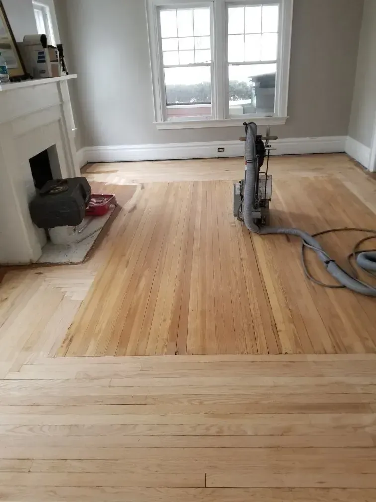 A wooden floor being sanded with a machine in a room. Fireplace and window in the background.