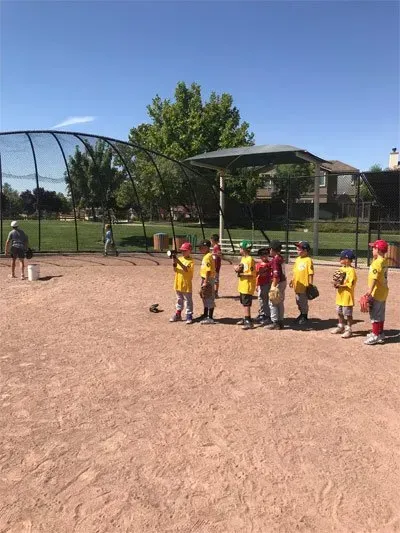 Children in yellow shirts stand near a baseball field backstop on a sunny day