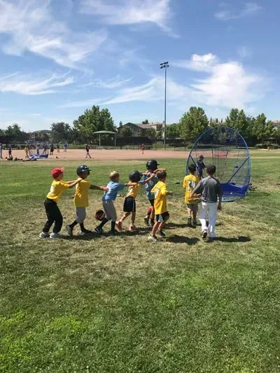 Children in yellow shirts line up at a soccer goal on a grassy field under a blue sky.