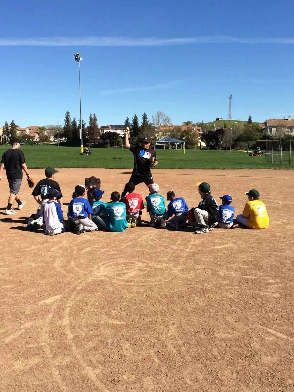 Children seated on a field watching a coach during an outdoor sports practice