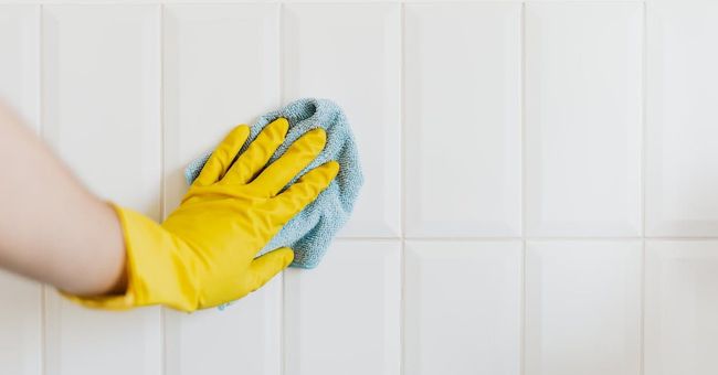 Een persoon met gele handschoenen maakt een witte tegelmuur schoon met een doek.