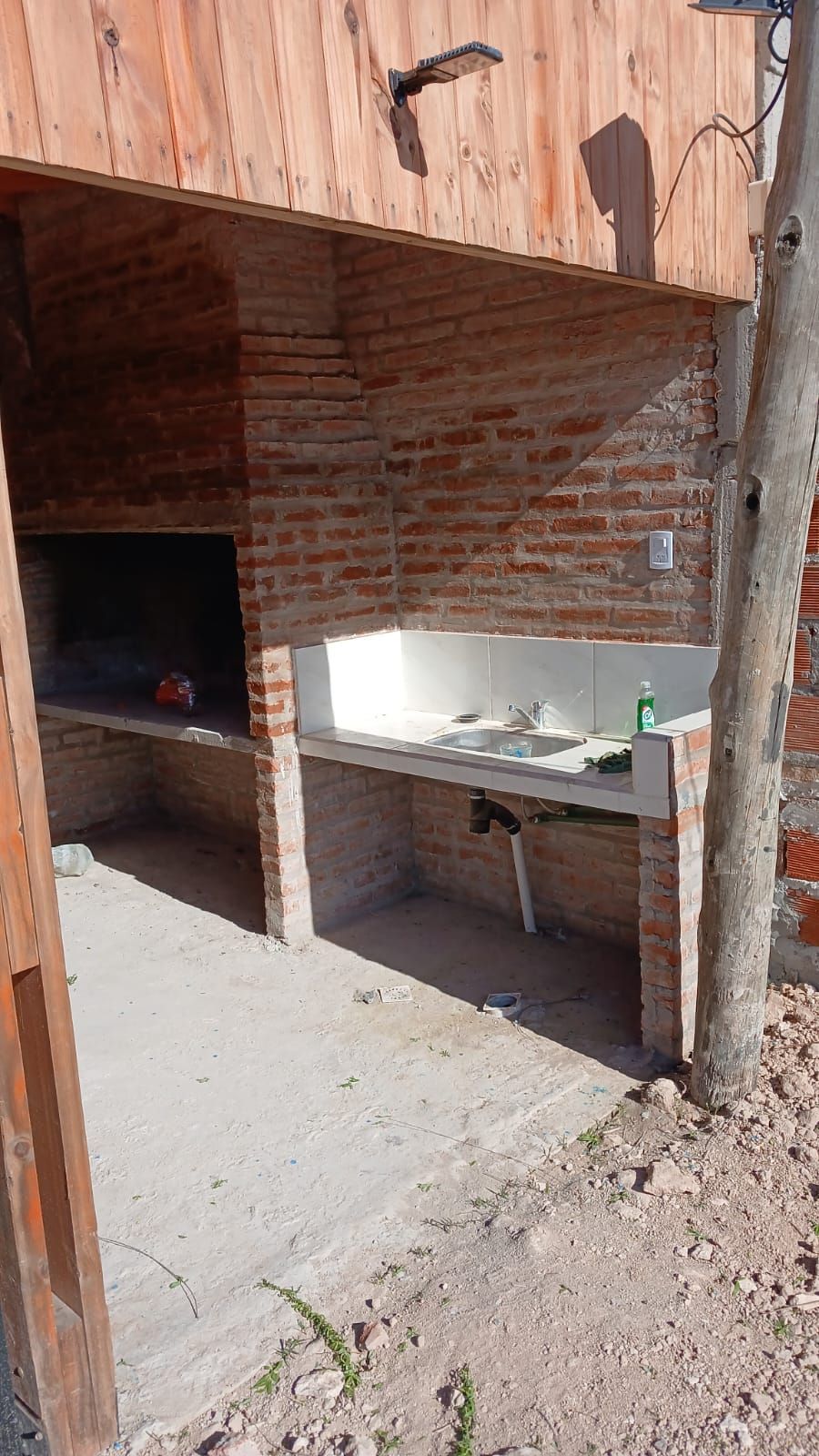 Zona de barbacoa de ladrillo al aire libre, con dos puestos de cocina abiertos y una encimera de hormigón bajo un techo de madera.