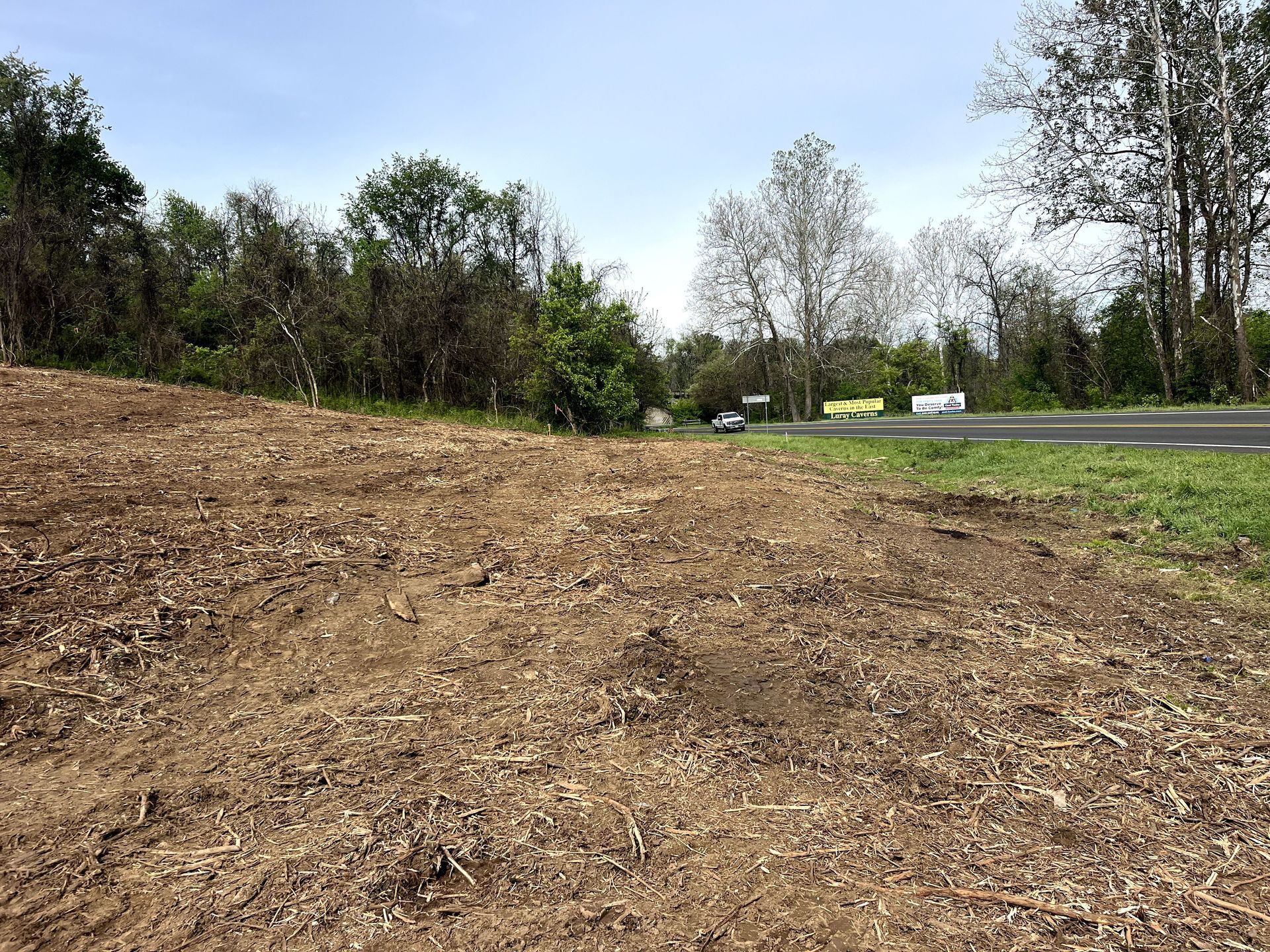Bare dirt roadside embankment beside a paved road with trees and cars in the background