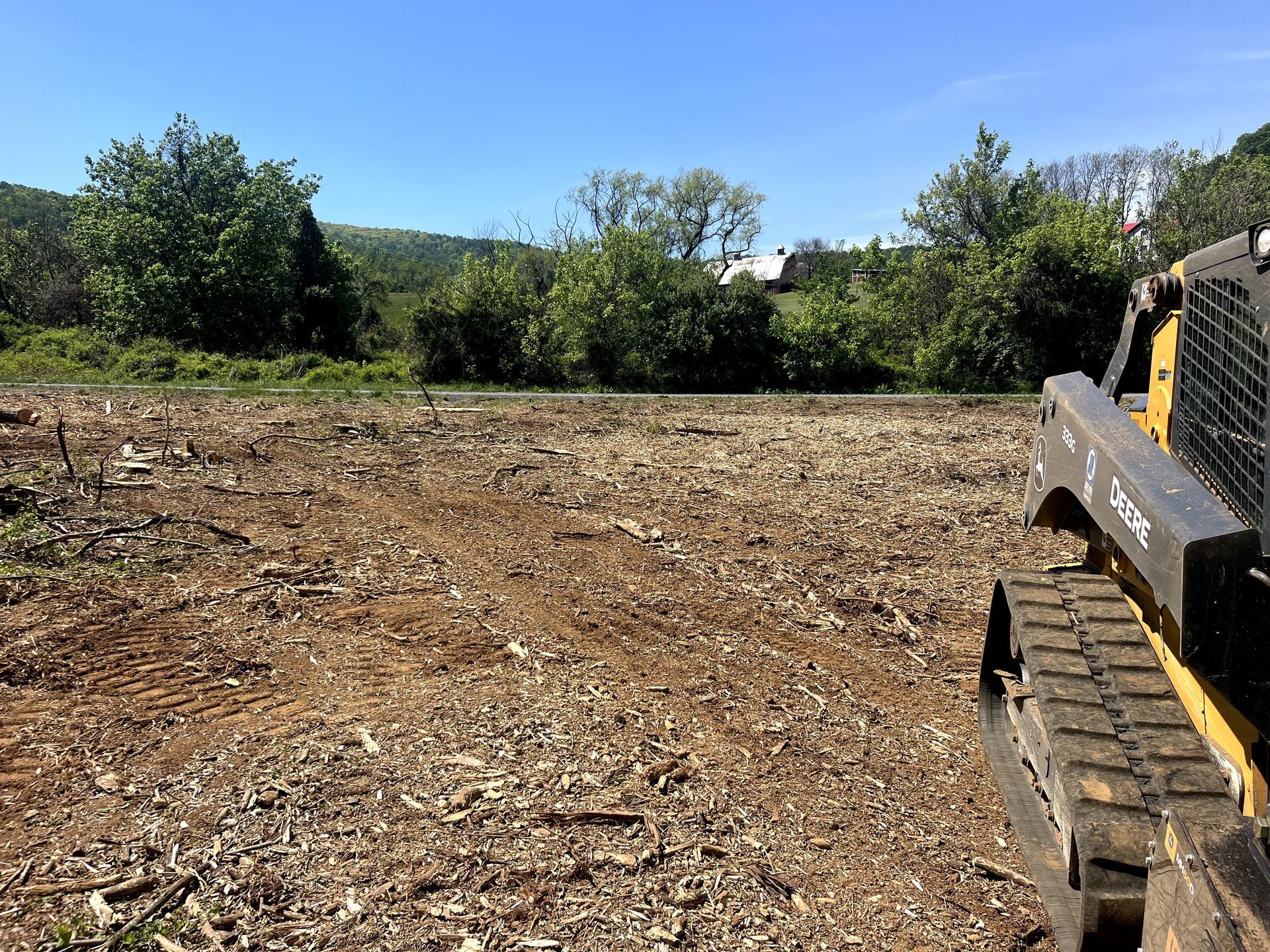 Cleared dirt lot with a skid-steer at right and trees lining the background under a blue sky