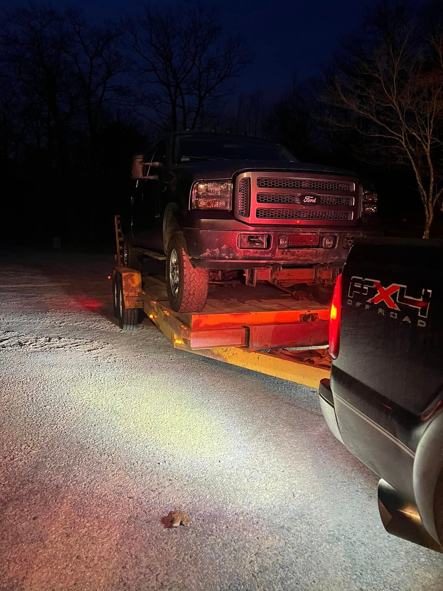 A black pickup truck towing another black pickup truck on a flatbed trailer at night.
