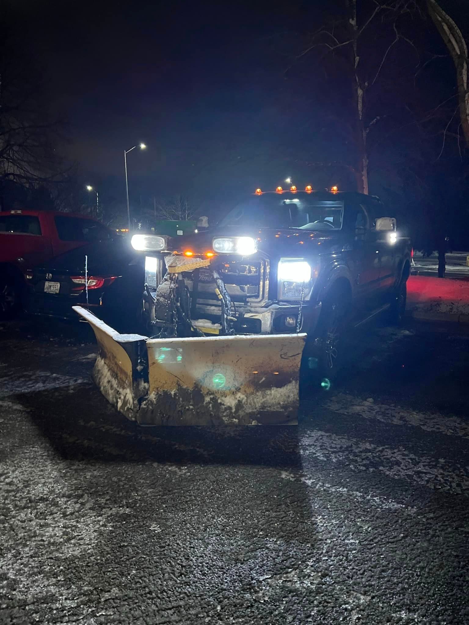 Snow plow truck clearing snow at night, headlights illuminated.